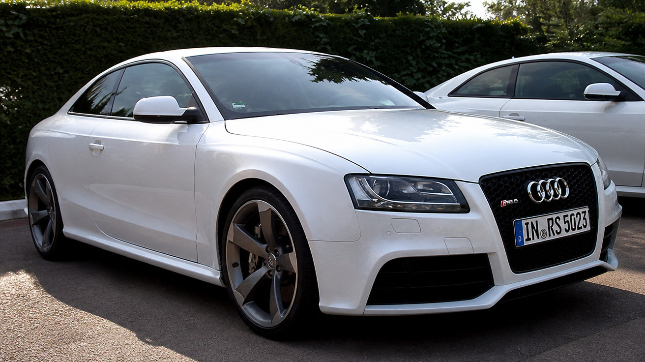 Weißer Audi RS5 Sportwagen, geparkt auf Asphalt vor einer grünen Hecke bei Tageslicht.