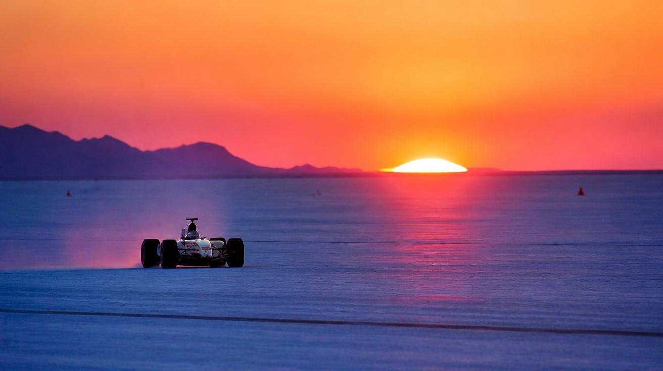 Formelrennwagen fährt bei Sonnenuntergang über eine weite, flache Salzfläche mit Bergen im Hintergrund.