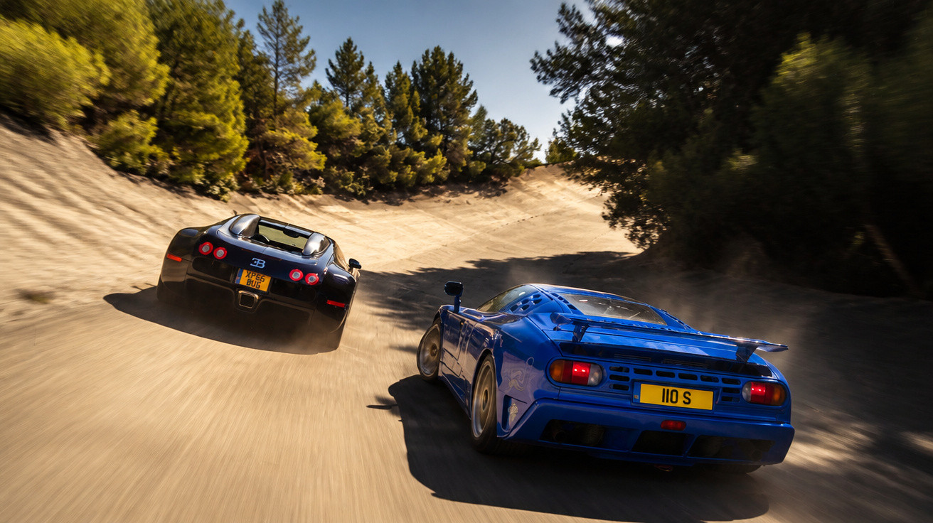 Zwei Sportwagen, ein schwarzer Bugatti und ein blauer Jaguar, fahren auf einer kurvigen Rennstrecke durch bewaldetes Gebiet.