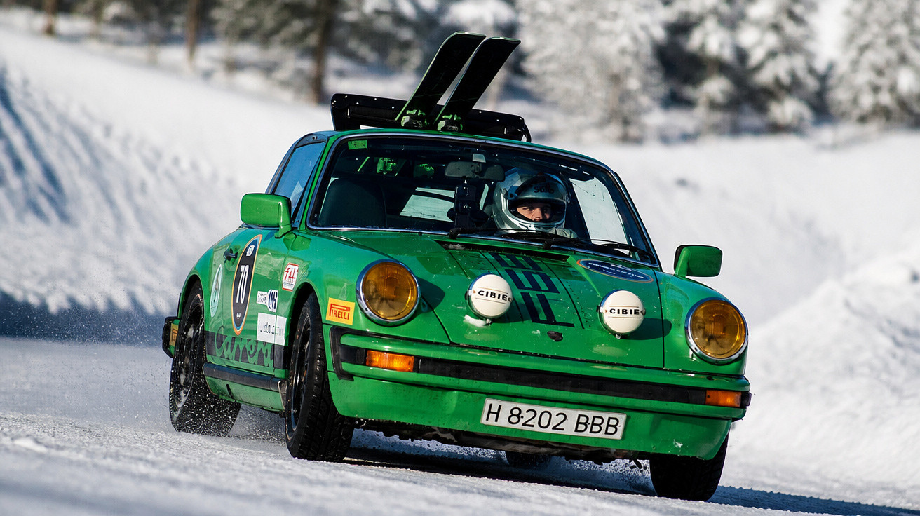 Grüner Porsche 911 mit Skiern auf Dach fährt im Schnee bei Winterrennen, Fahrer mit Helm sitzt am Steuer.