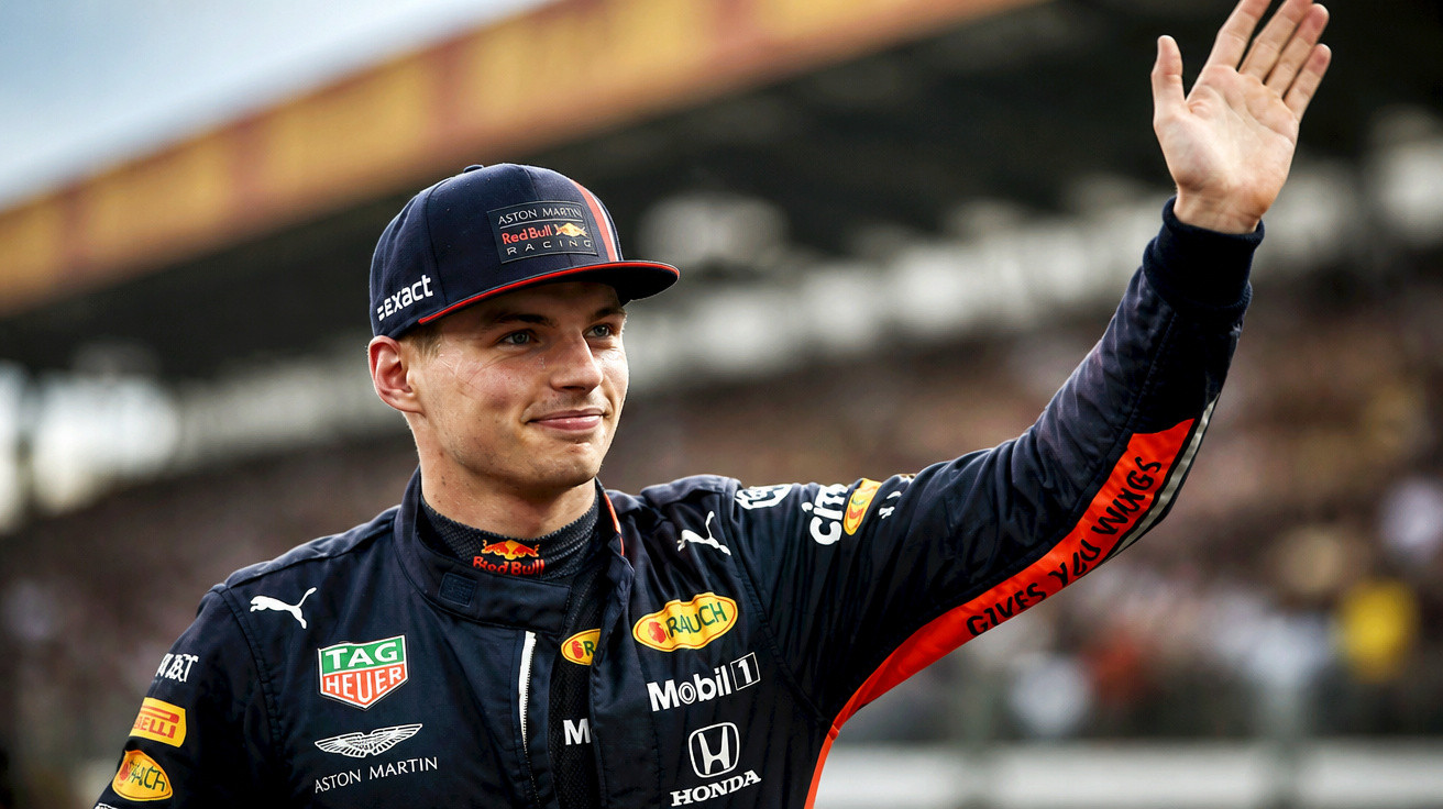 Formel-1-Fahrer Max Verstappen in Rennanzug und Cap hebt lächelnd die Hand vor unscharfer Kulisse.