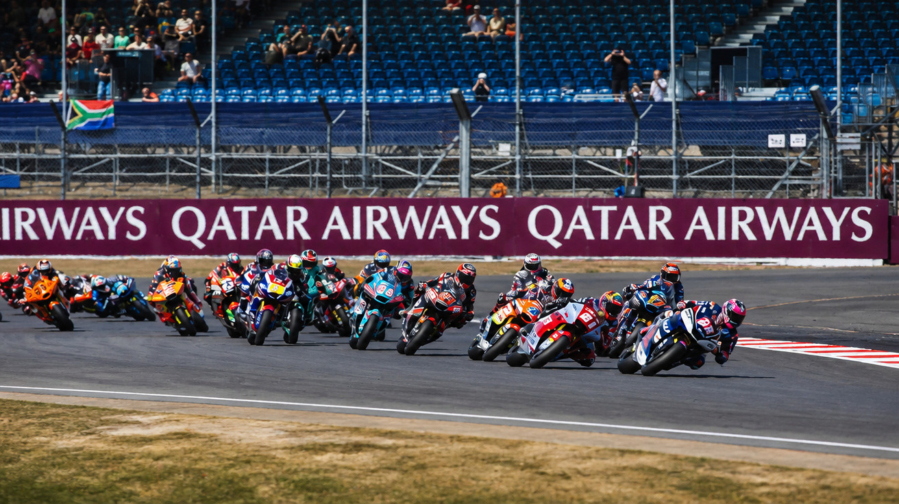 Motorradrennen mit mehreren Fahrern in Kurvenlage auf Rennstrecke vor vollen Tribünen und Qatar Airways Werbung.