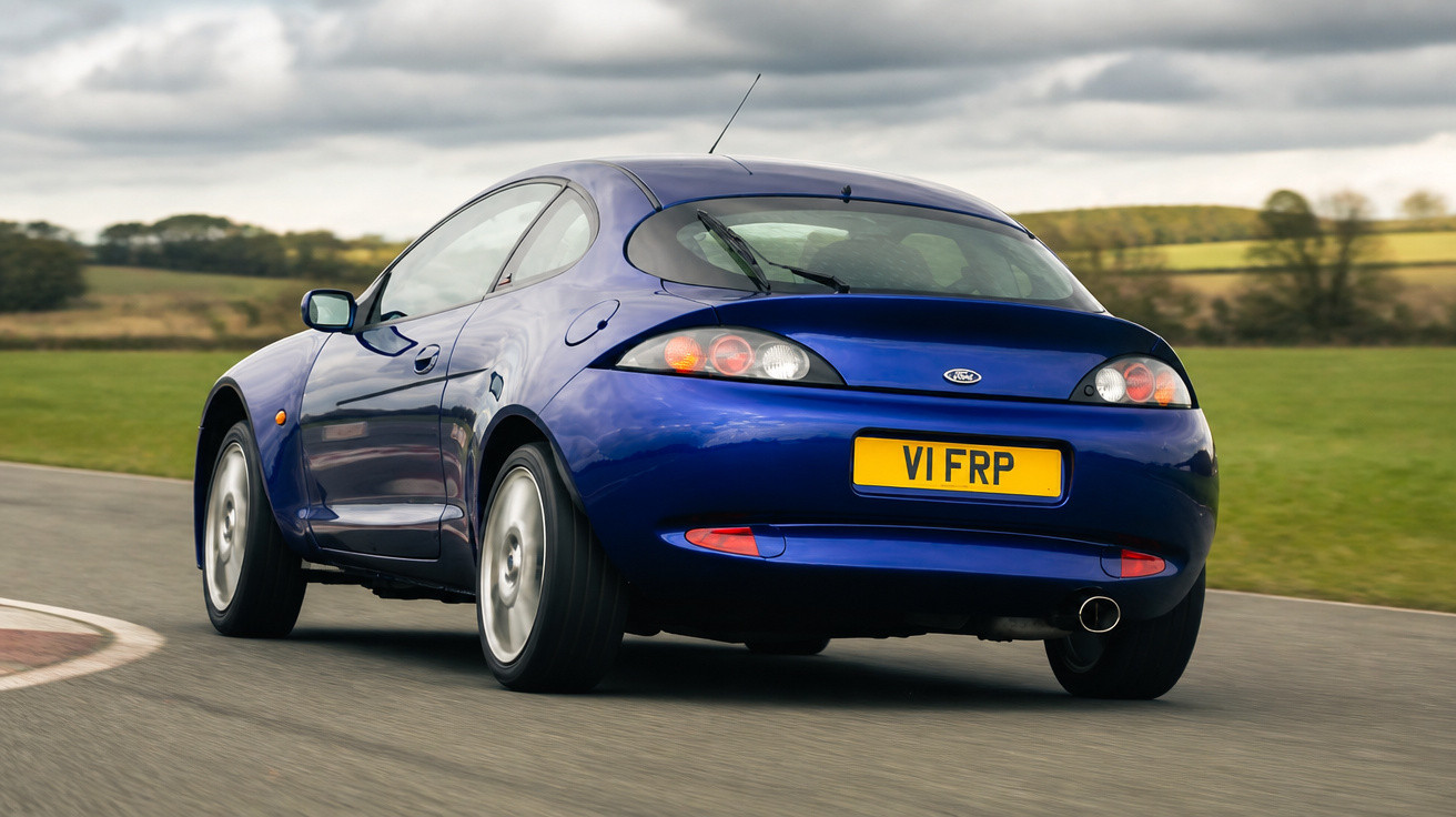 Blauer Ford Puma auf Rennstrecke, Rückansicht, an bewölktem Tag, umgeben von grüner Landschaft.